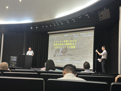 ホール形式の講演会場で行われているシンポジウムの様子。前方のスクリーンには「ミャンマー中部における後期中新世の環境変化と化石ホミノイドの盛衰」と題した発表スライドが投影され、登壇者が説明を行っている。客席には参加者が着席している。