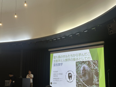 講演会場の大型スクリーンに「屋久島のサルたちから学んだこと：生態学と人類学の接点としての霊長類学」と題した発表スライドが映し出されている。演壇では発表者がマイクを持って講演し、スライドにはサルの写真と研究機関のロゴが表示されている。